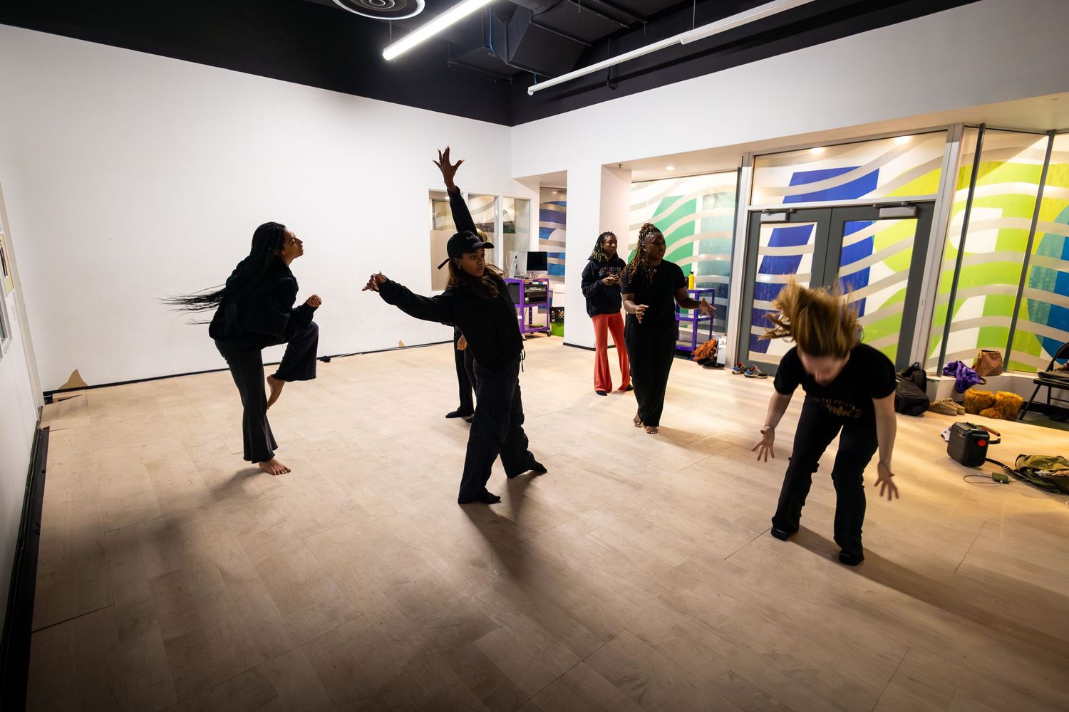 Group of people practicing dance moves in a modern studio with wood floors and colorful artwork on walls.