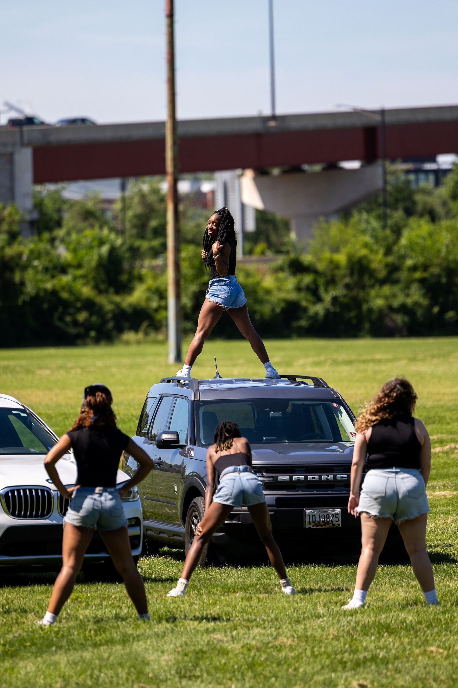 Four women in denim shorts dance outdoors, one standing on a Ford Bronco roof, with a highway overpass in the background.