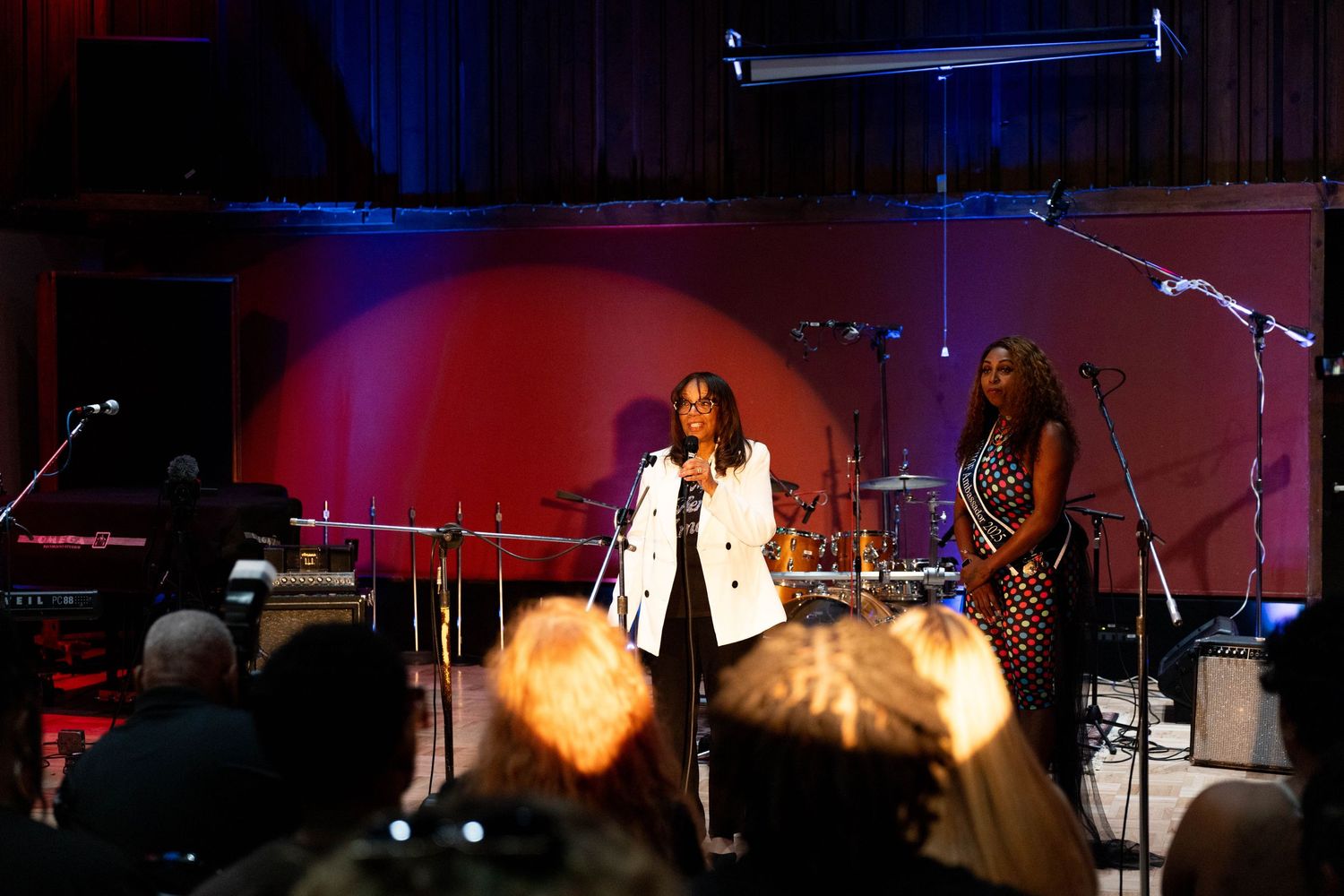 Two women performing on stage at a live music event, with red and pink stage lighting in the background.