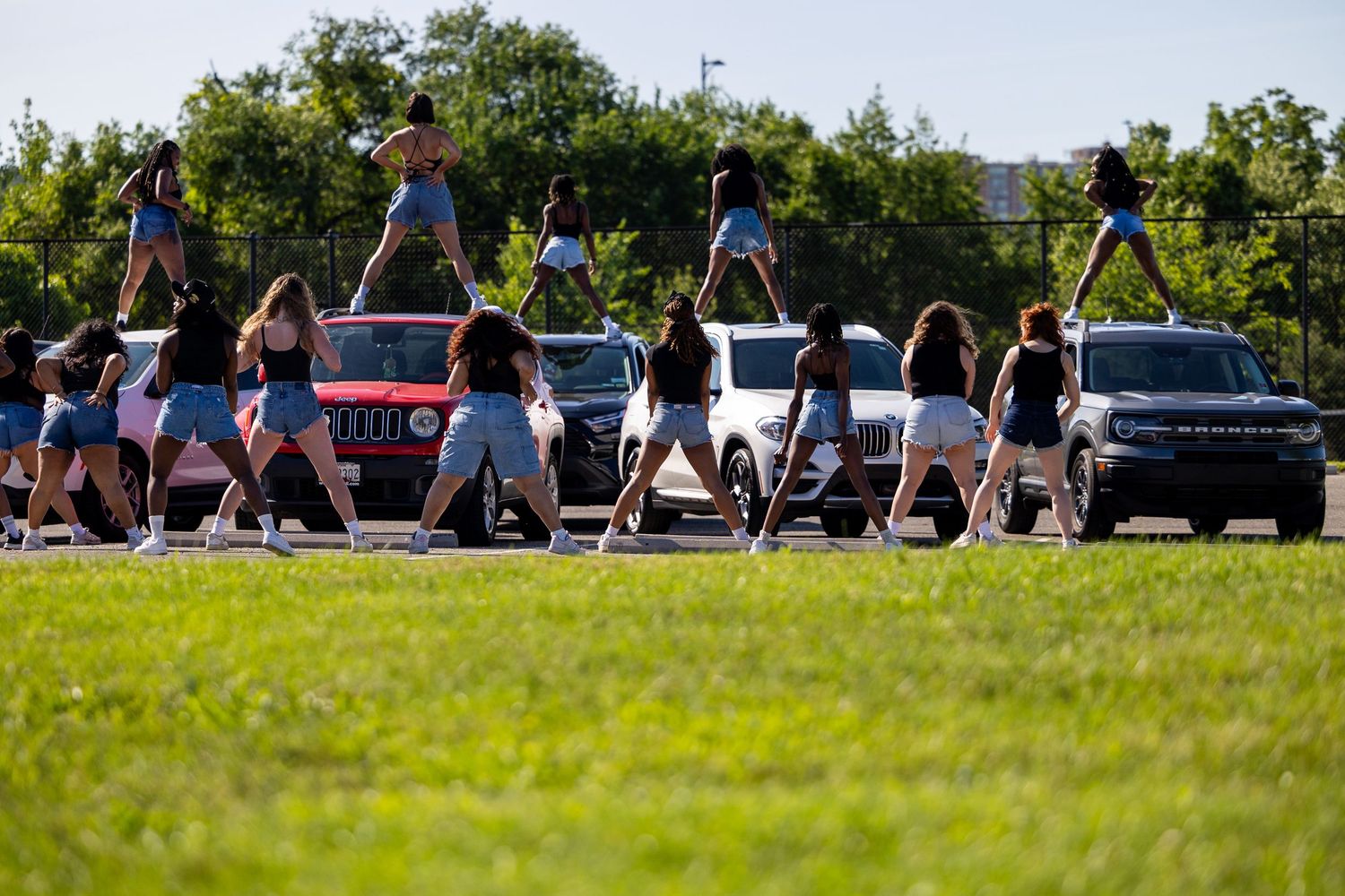 Cheerleaders performing synchronized dance routine outdoors near parked cars on a sunny day.