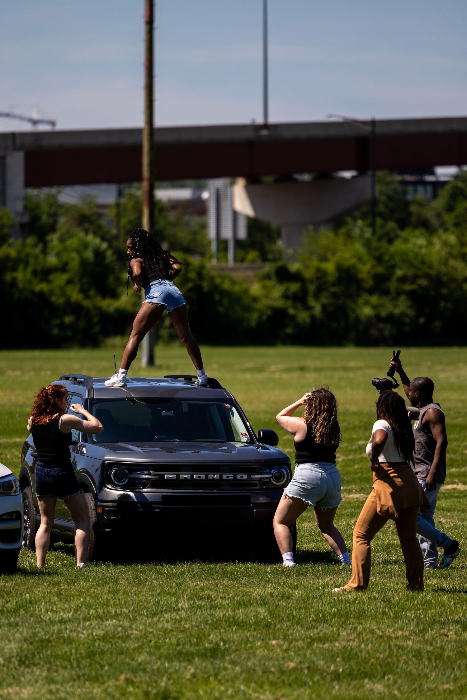 Woman standing on top of a Ford Bronco SUV on grass while a photographer and group of people gather around.