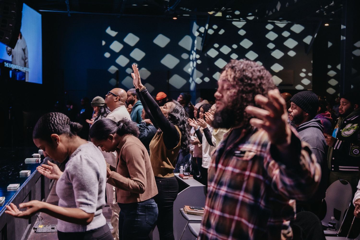 Concert crowd raises hands in worship during an emotional performance at a dimly lit venue.