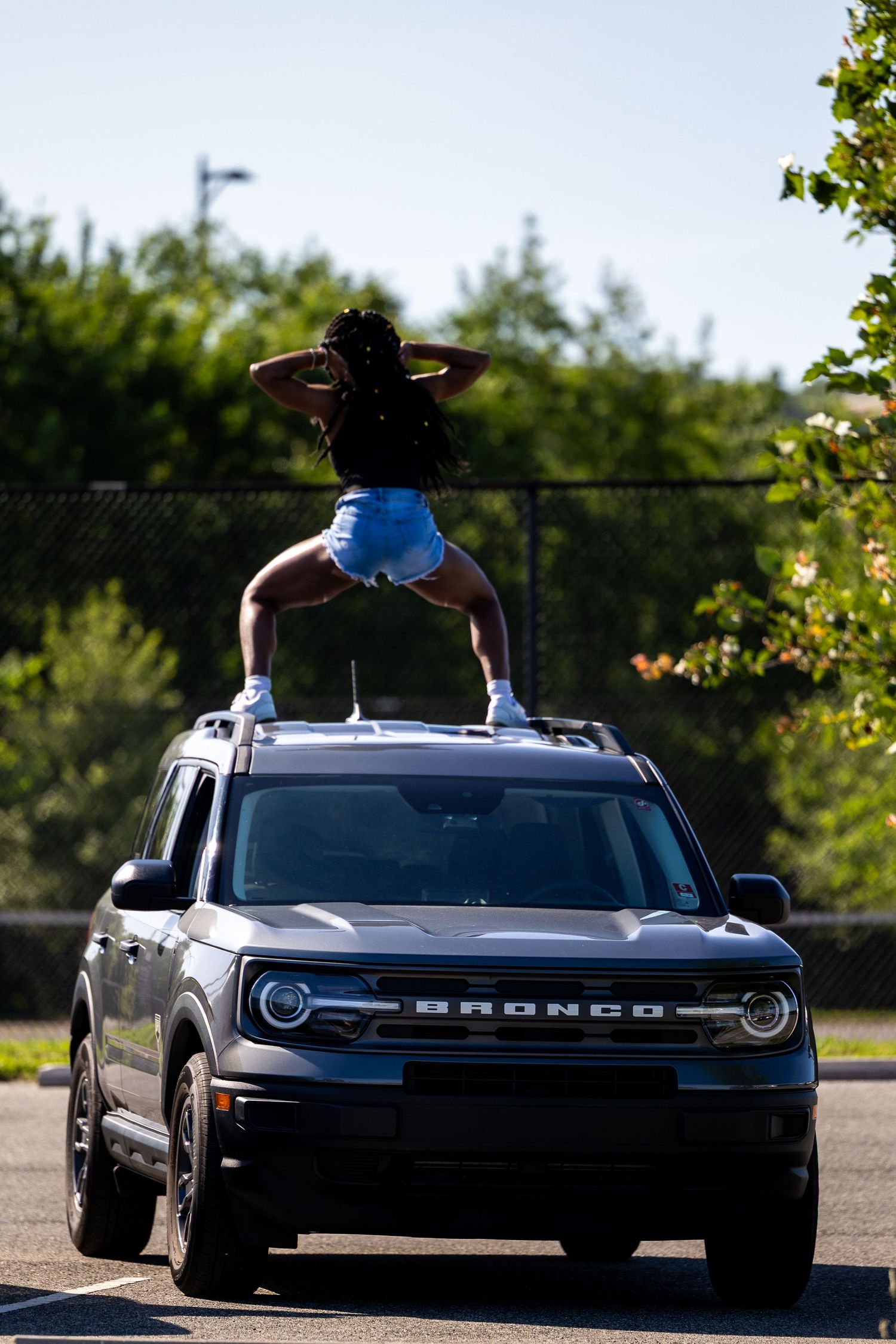 Person standing on top of a Ford Bronco SUV in a parking lot with trees in the background.