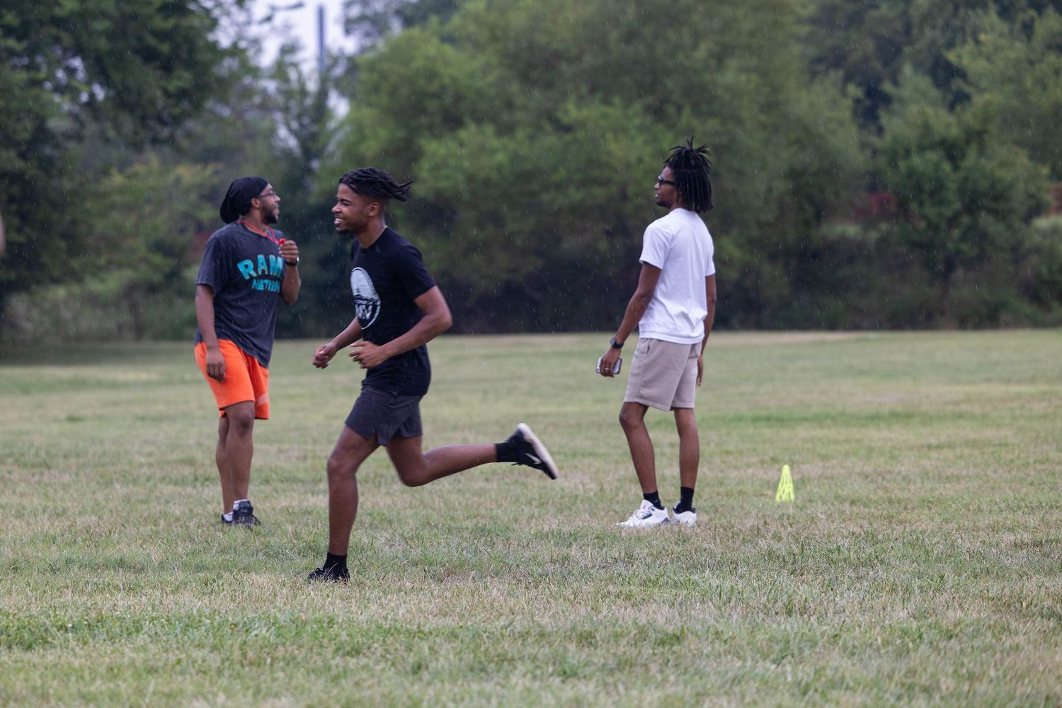 Young athletes run and train on a grassy field outdoors, with a yellow cone marking the course.