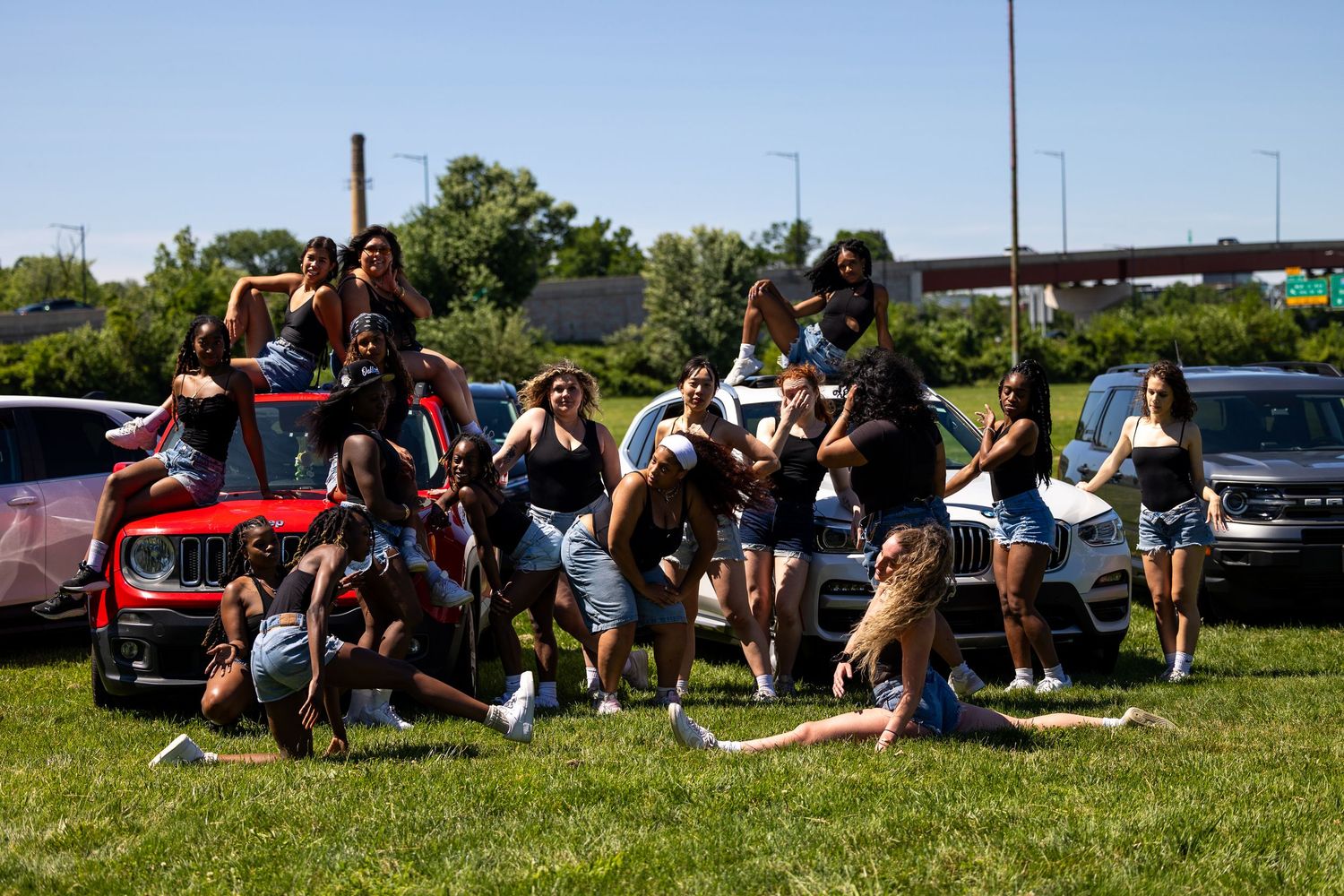 A large group of young women posing and dancing together on grass in a parking lot on a sunny day.