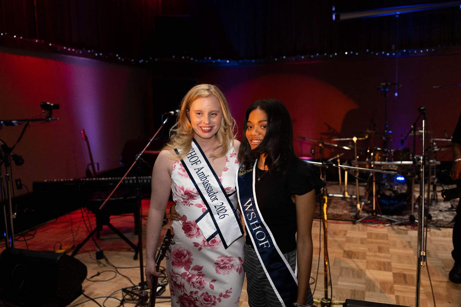 Two beauty pageant contestants wearing sashes pose together smiling at an indoor event with stage lighting.