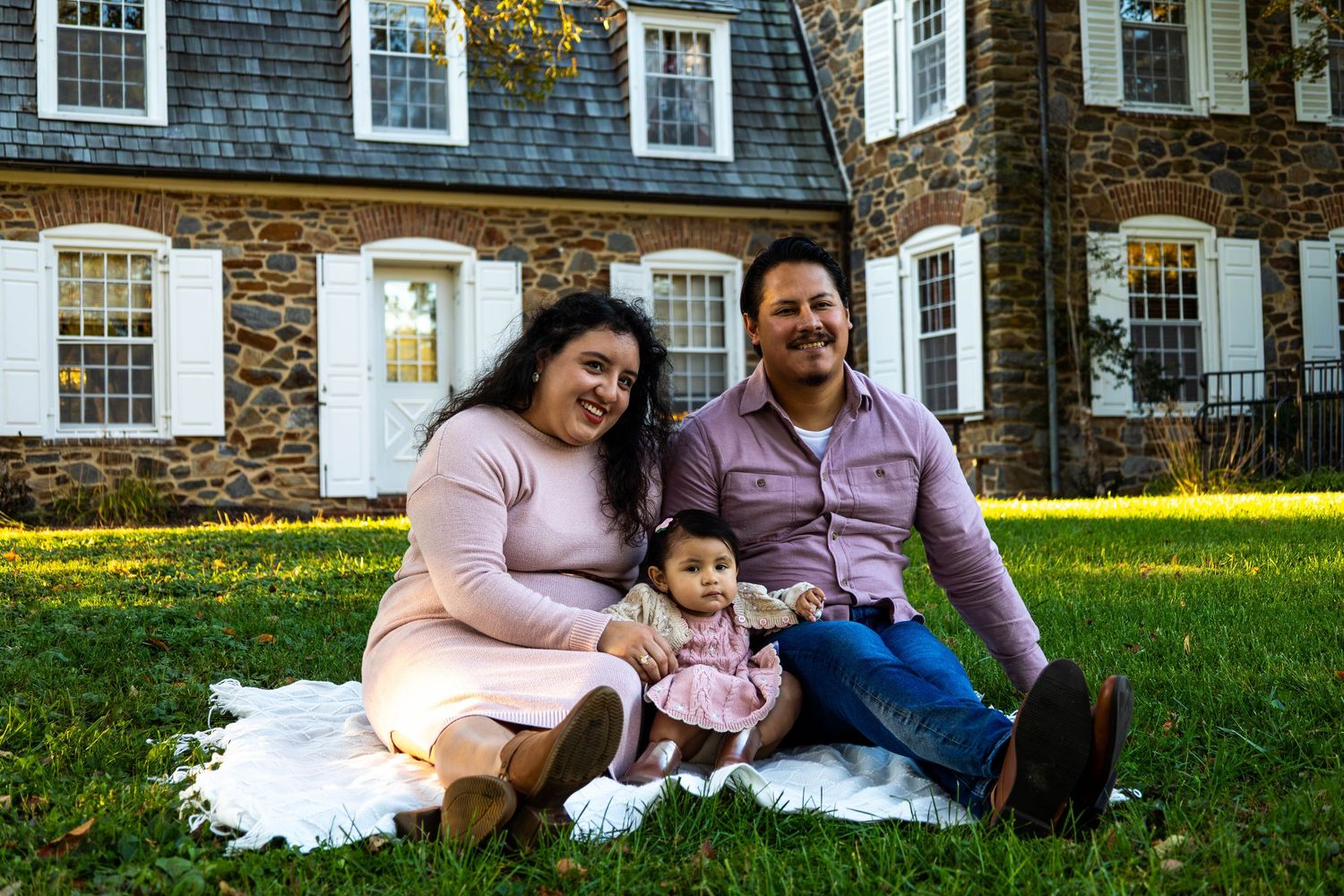Family of three sitting on white blanket on grass in front of stone house, smiling at camera.