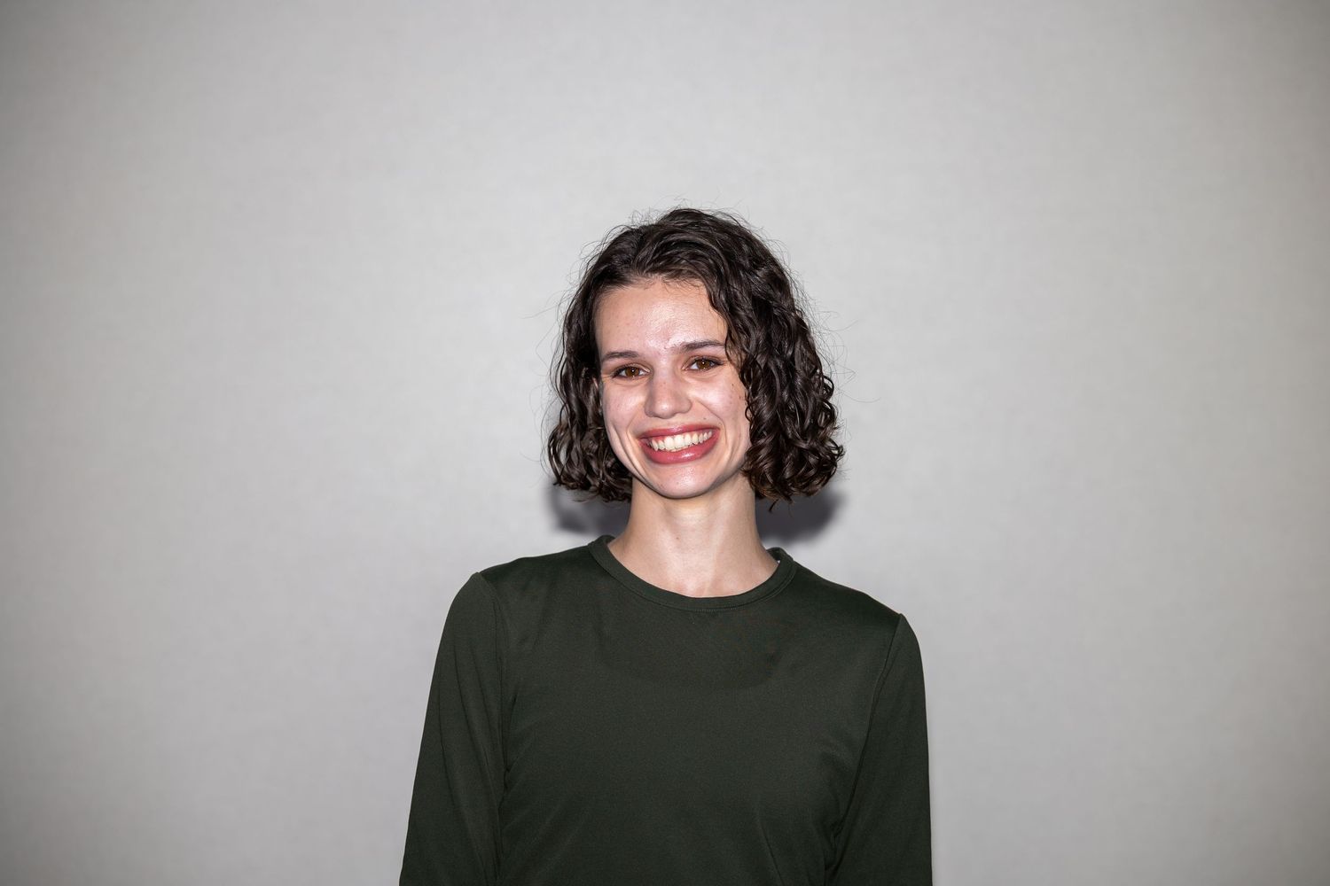Smiling young woman with short curly dark hair wearing a dark green long-sleeve shirt against a gray background.