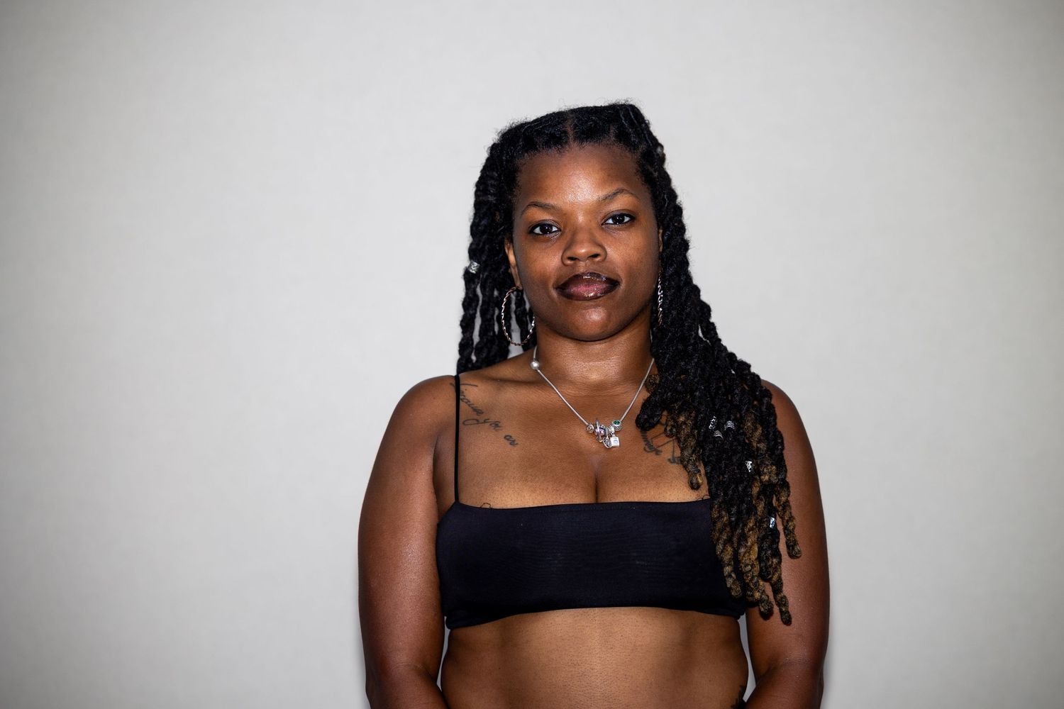 A woman with braided locs wearing a black bandeau top and silver necklace poses against a light gray background.