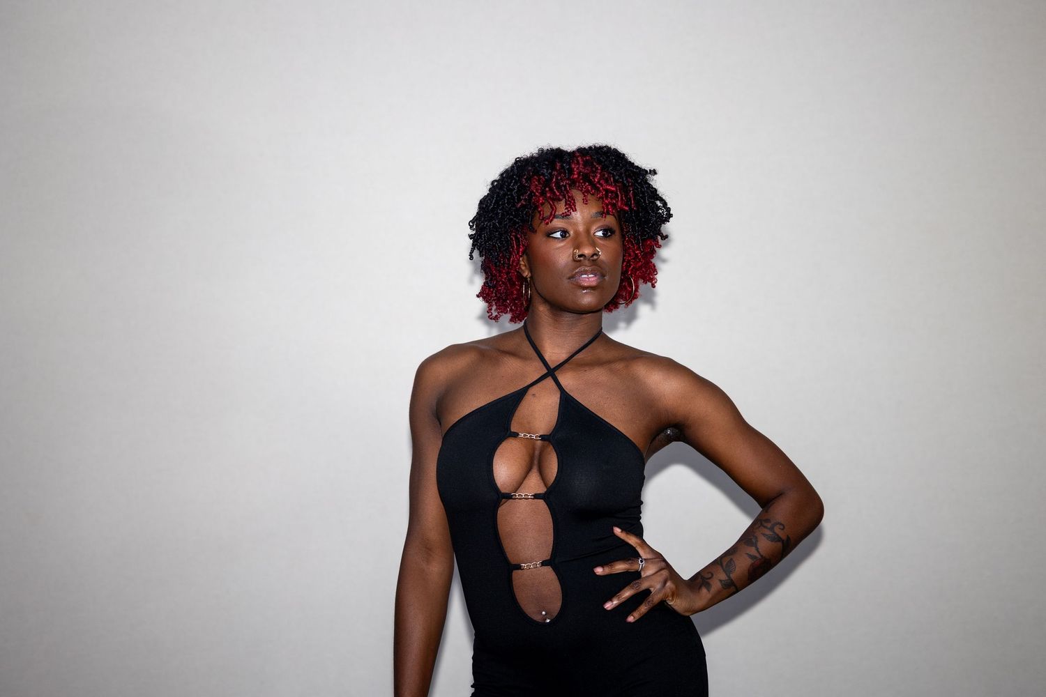 Confident woman with red curly hair in a black cutout halter dress, posing with one hand on hip.