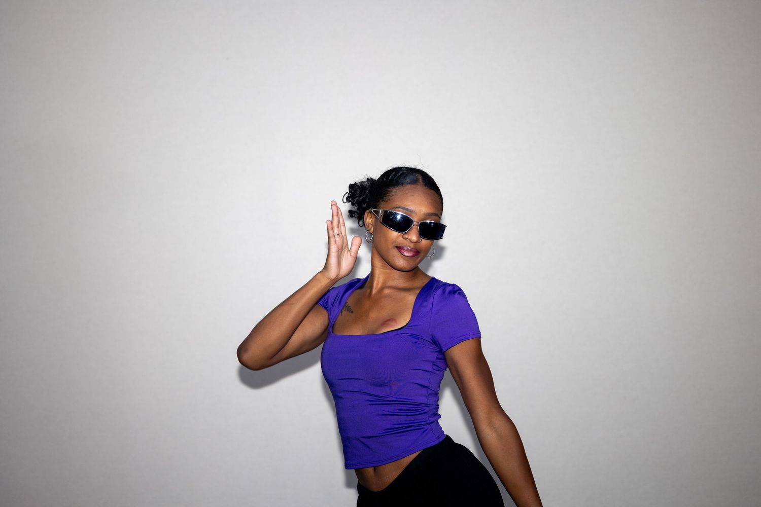 Stylish young woman in purple crop top and sunglasses posing with hand raised against white background.