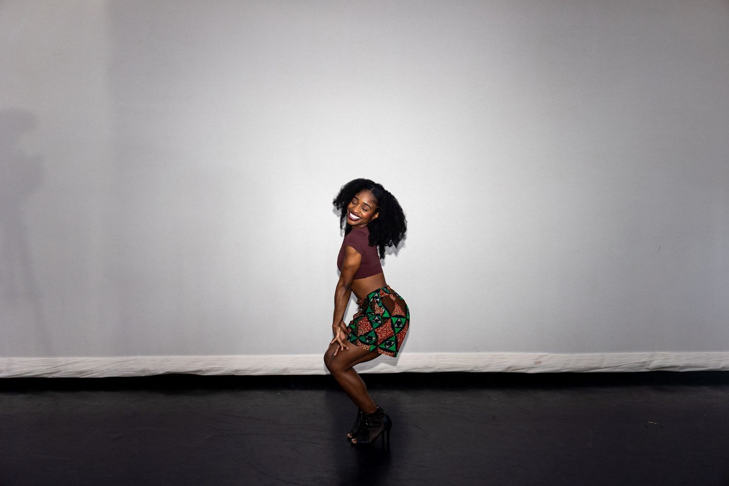 Woman in vibrant African print skirt posing gracefully in profile against a neutral gray background.