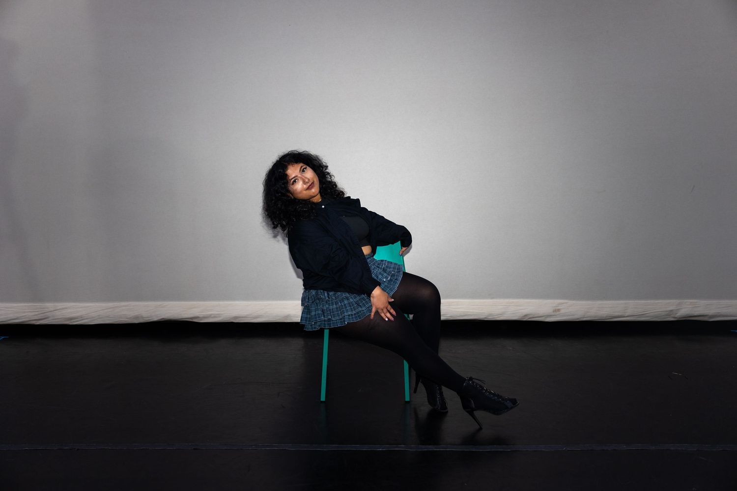 Woman in blue skirt and black sweater sitting on green chair against gray background.