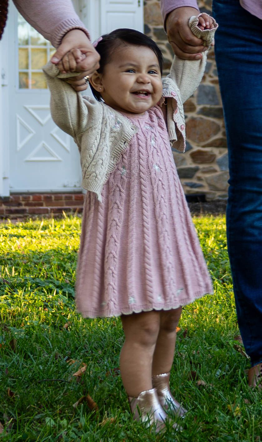 Smiling toddler girl in pink knit dress and cream cardigan stands on green grass holding adults hands.