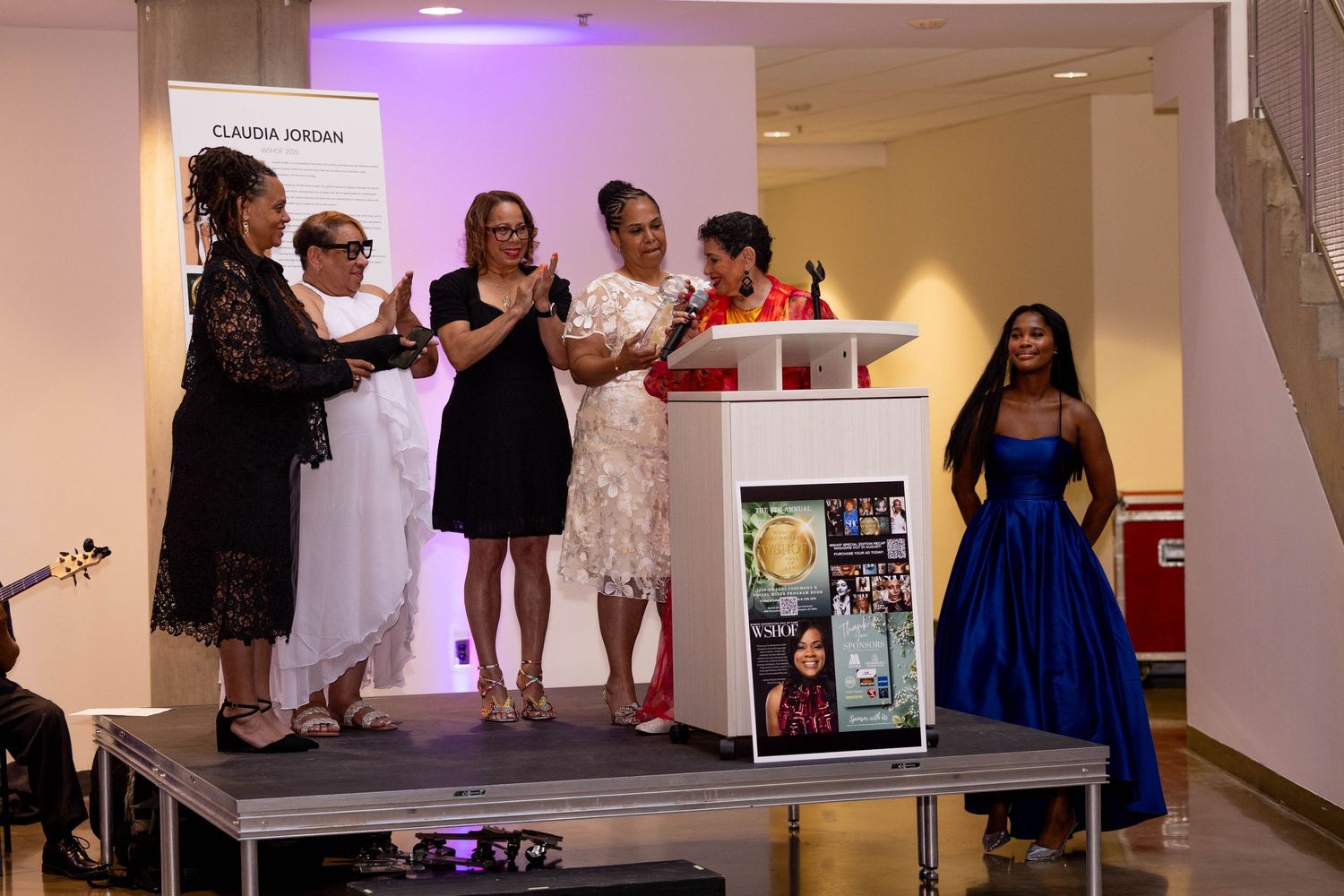 Group of women celebrate on stage at Claudia Jordan event, including one in a bright blue dress.
