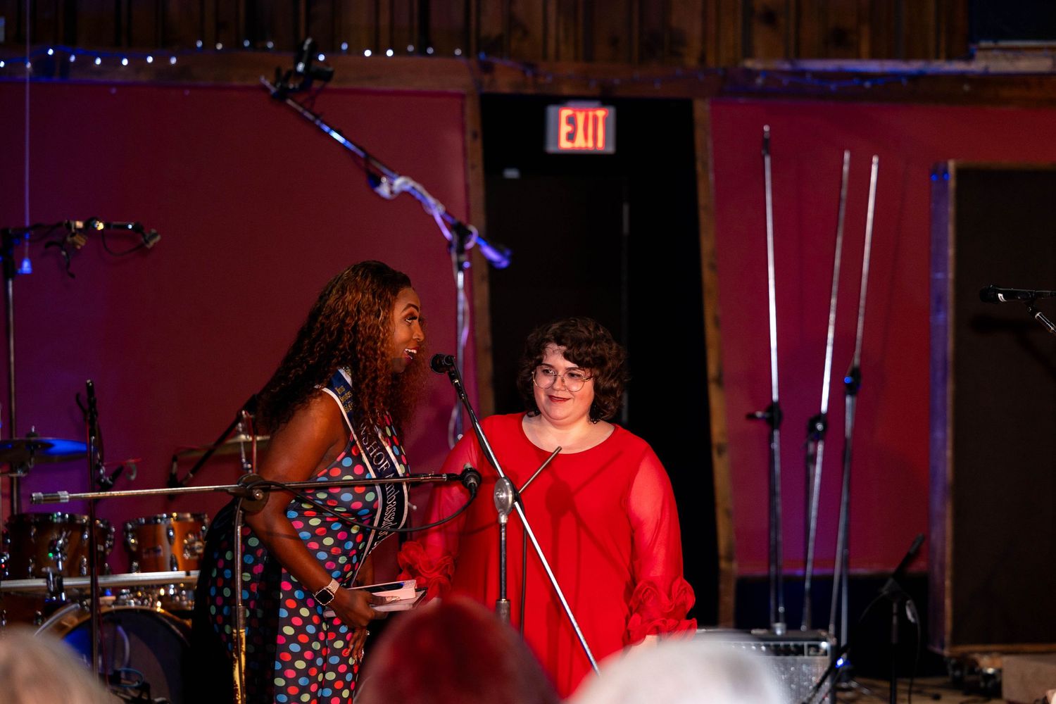 Two women on stage at a live music venue, one in a patterned dress and one in red, speaking at microphones.