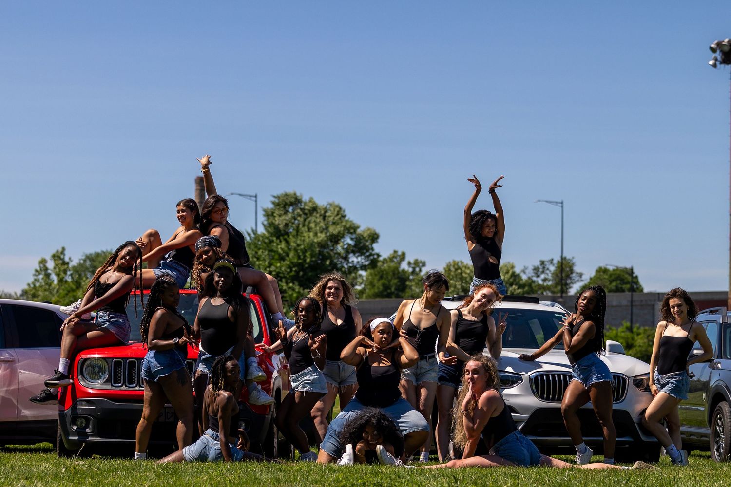 Large group of young women posing together outdoors near cars on a sunny day.