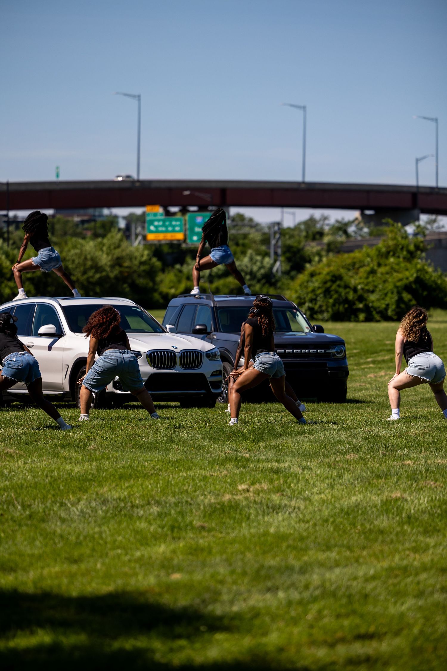 Women in denim shorts dancing near luxury SUVs on green grass near a highway overpass on a sunny day.