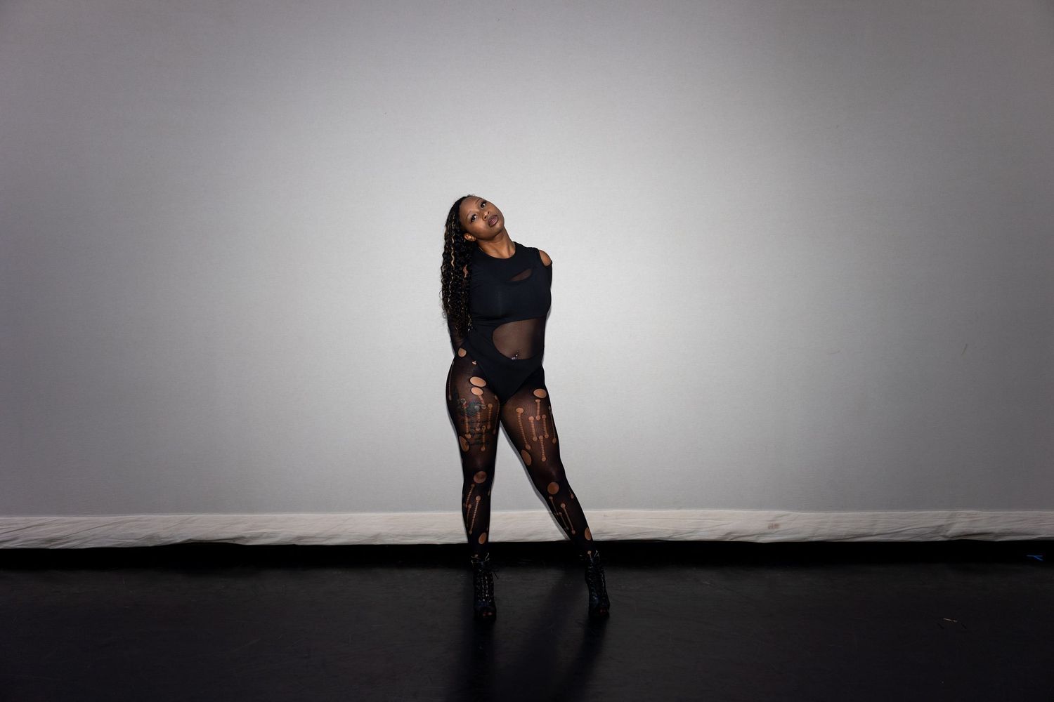 A dancer in a sheer black bodysuit and heels performs an expressive lean in a gray dance studio.