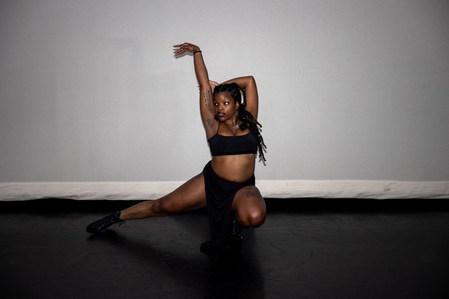 Black female dancer in black outfit performing a dramatic low lunge pose on a dark studio floor.