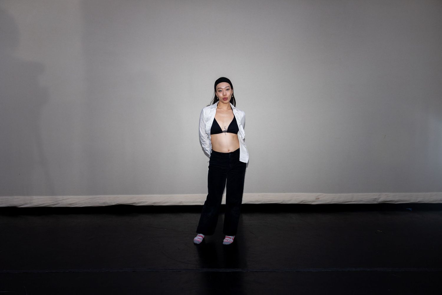 Woman poses in white blazer, black bra, and trousers on dark studio floor.