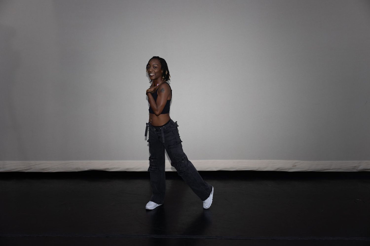 Slender woman in dark cargo pants and brown crop top walks confidently in a studio space.