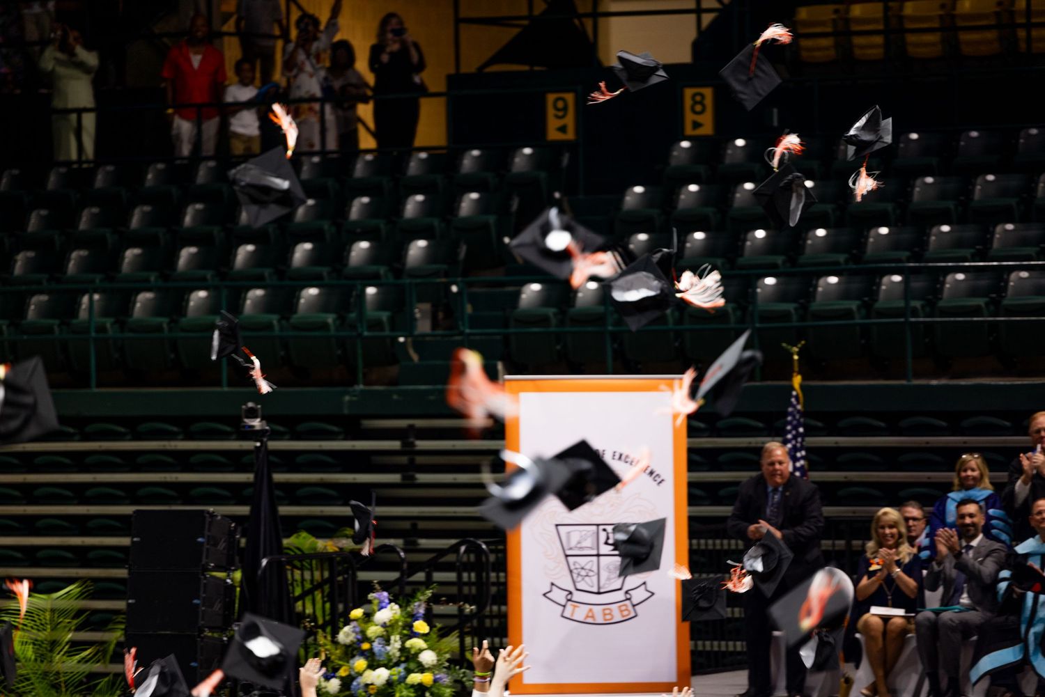 Graduation caps tossed in the air during a university commencement ceremony inside an indoor arena.