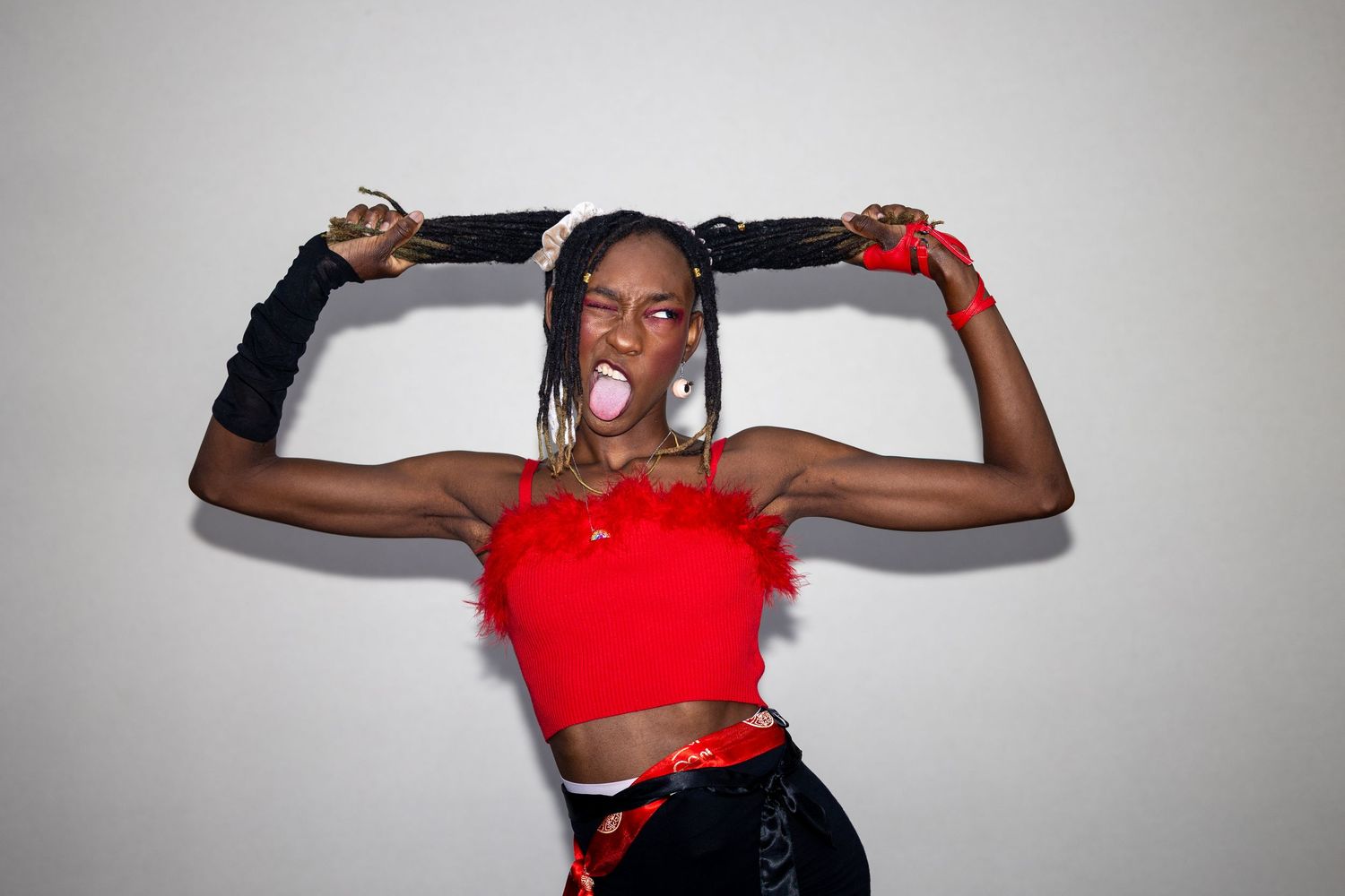 A muscular Black woman in a red feather crop top holds her braids up while blowing a pink bubble gum bubble.