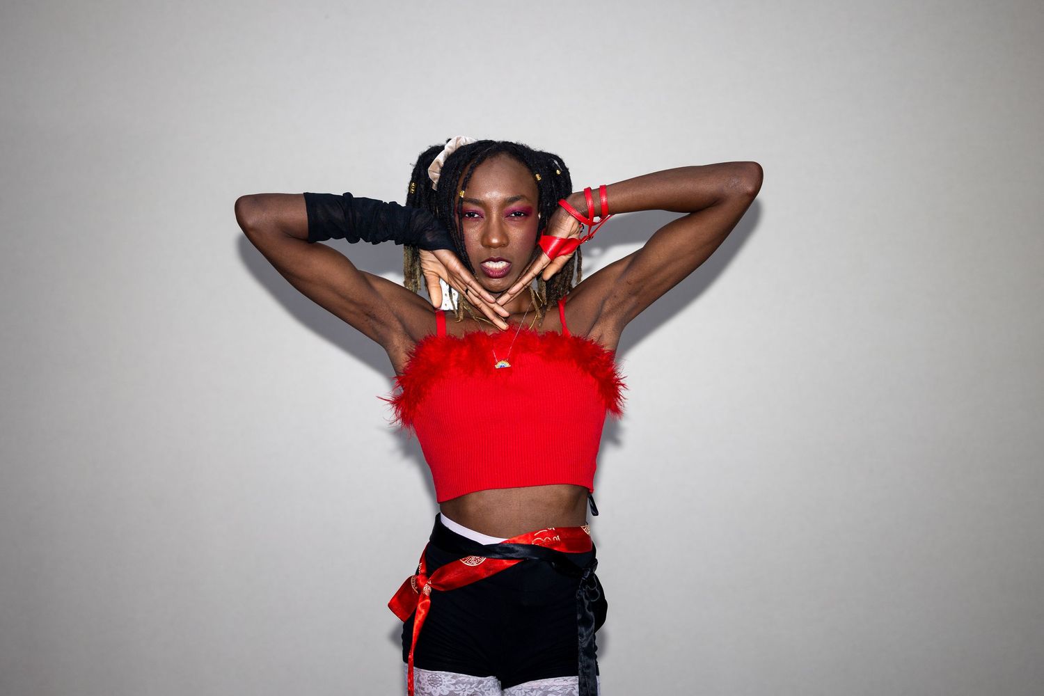 A Black woman in a red feathered crop top and black gloves strikes a bold pose with arms raised behind her head.