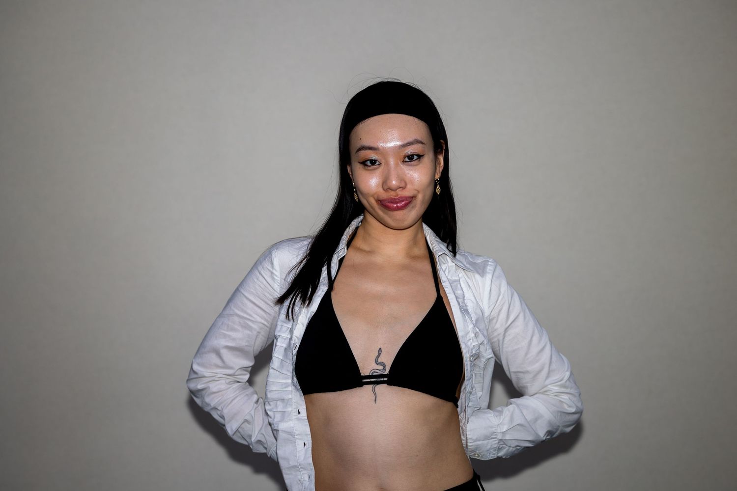 Young woman in black bikini top and open white jacket posing confidently against a gray wall.