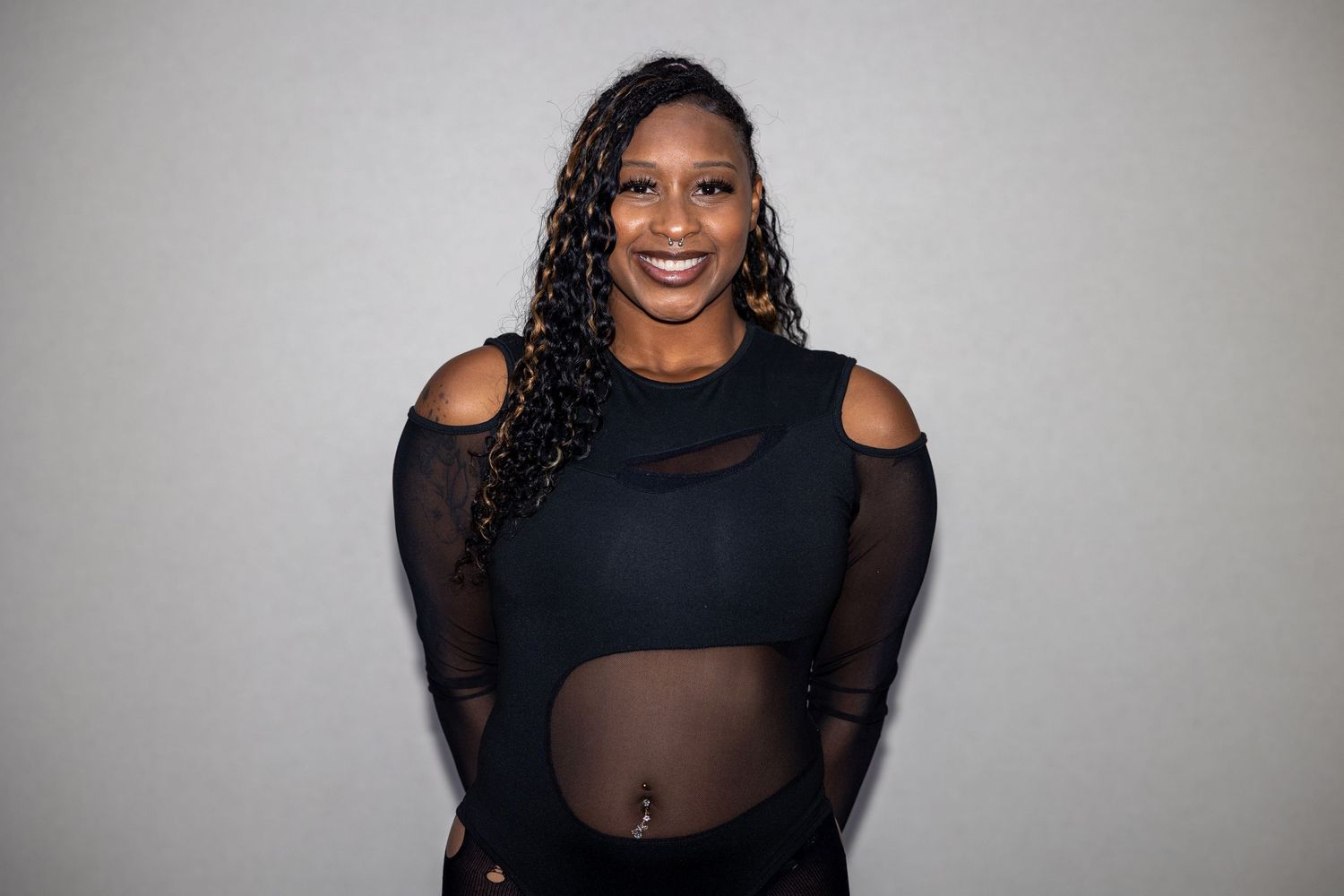Smiling woman in black sheer mesh bodysuit poses confidently against light gray background.