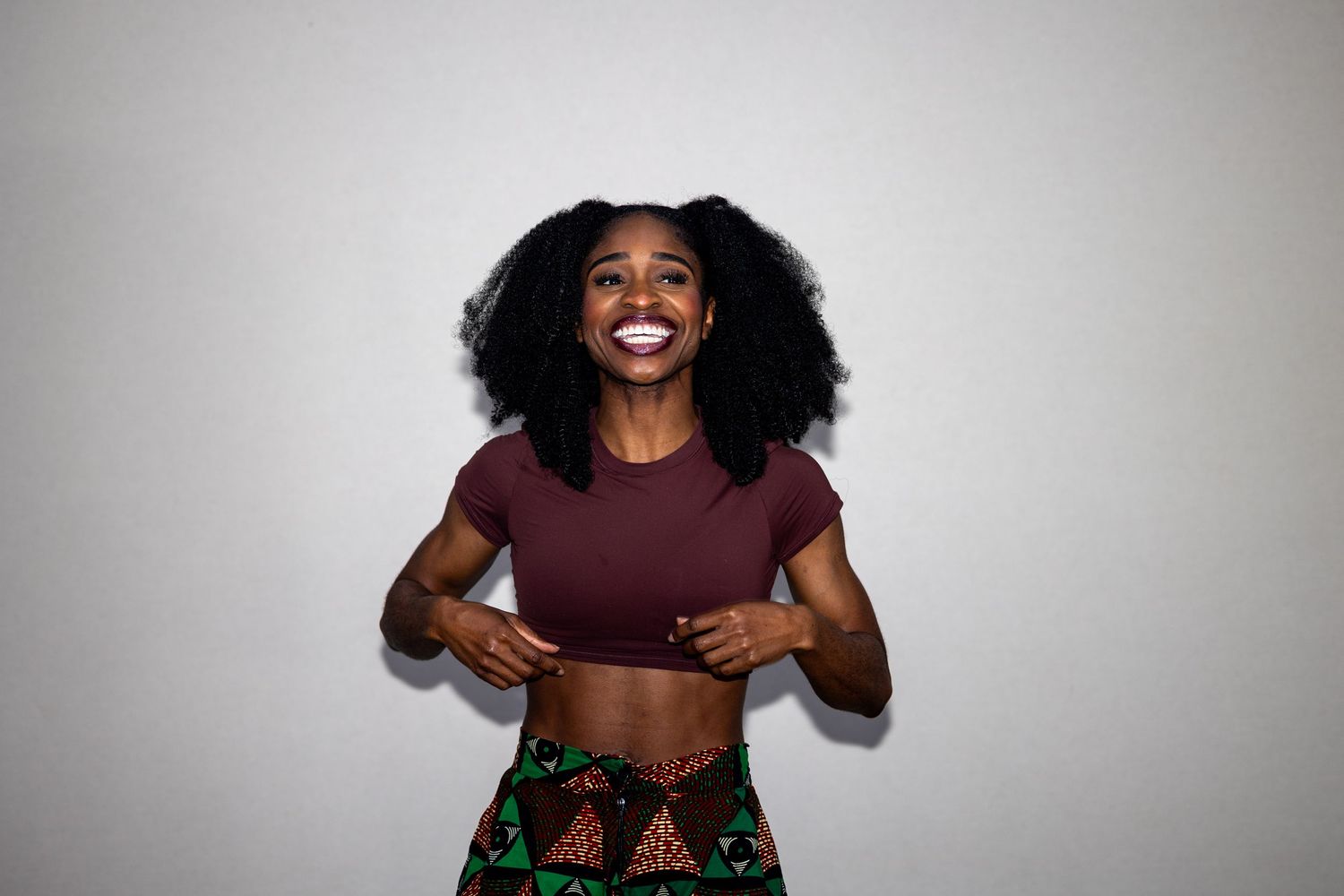 Joyful young woman with natural afro laughing in a maroon crop top and vibrant printed skirt.