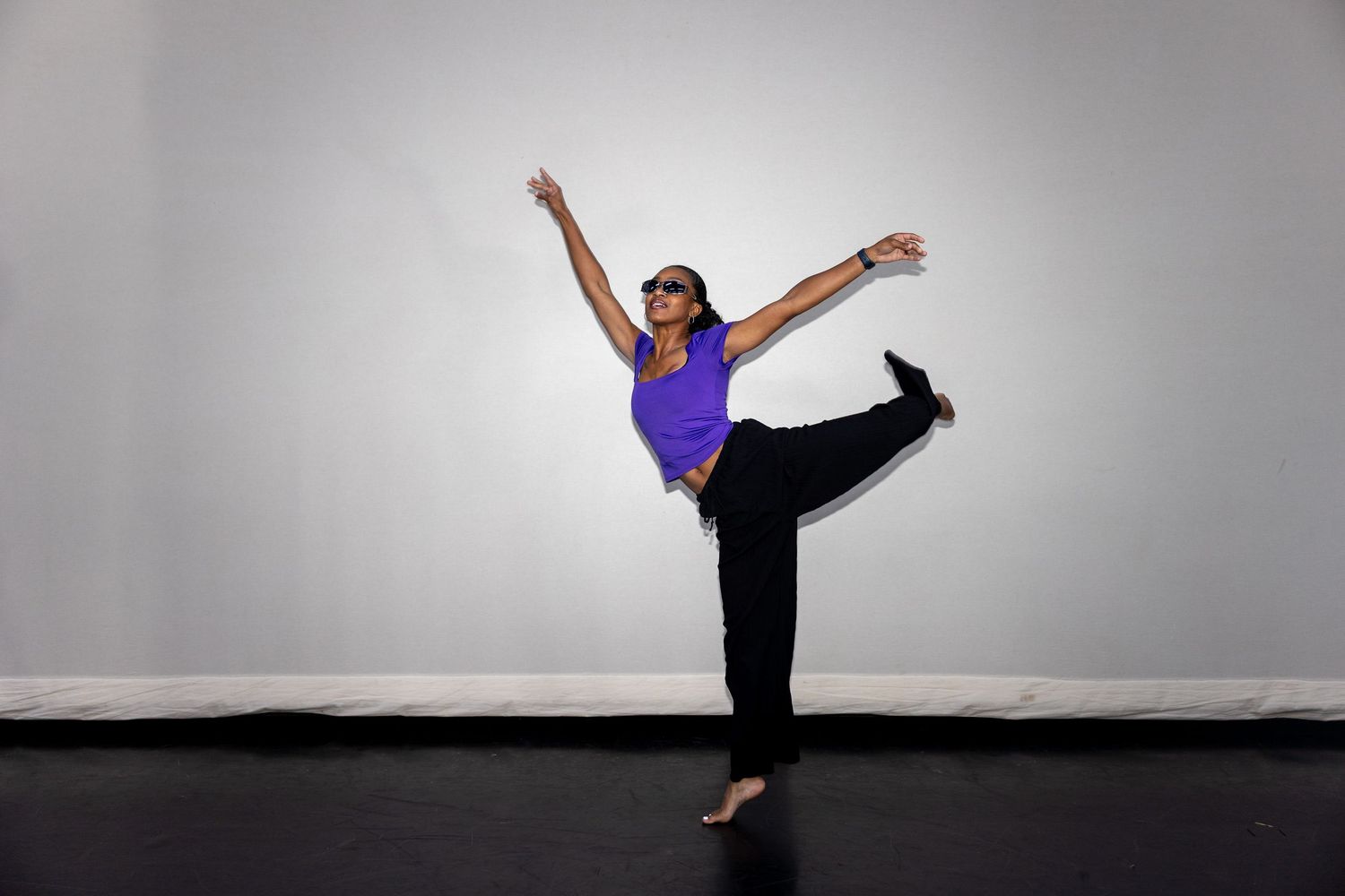 Woman in purple top and black pants performs a graceful ballet pose on one leg with arms extended in a studio.