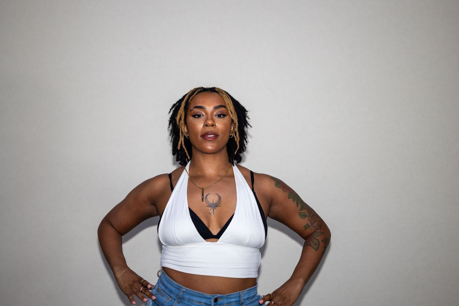 A confident tattooed woman in a white halter top and jeans poses with hands on hips against a gray background.