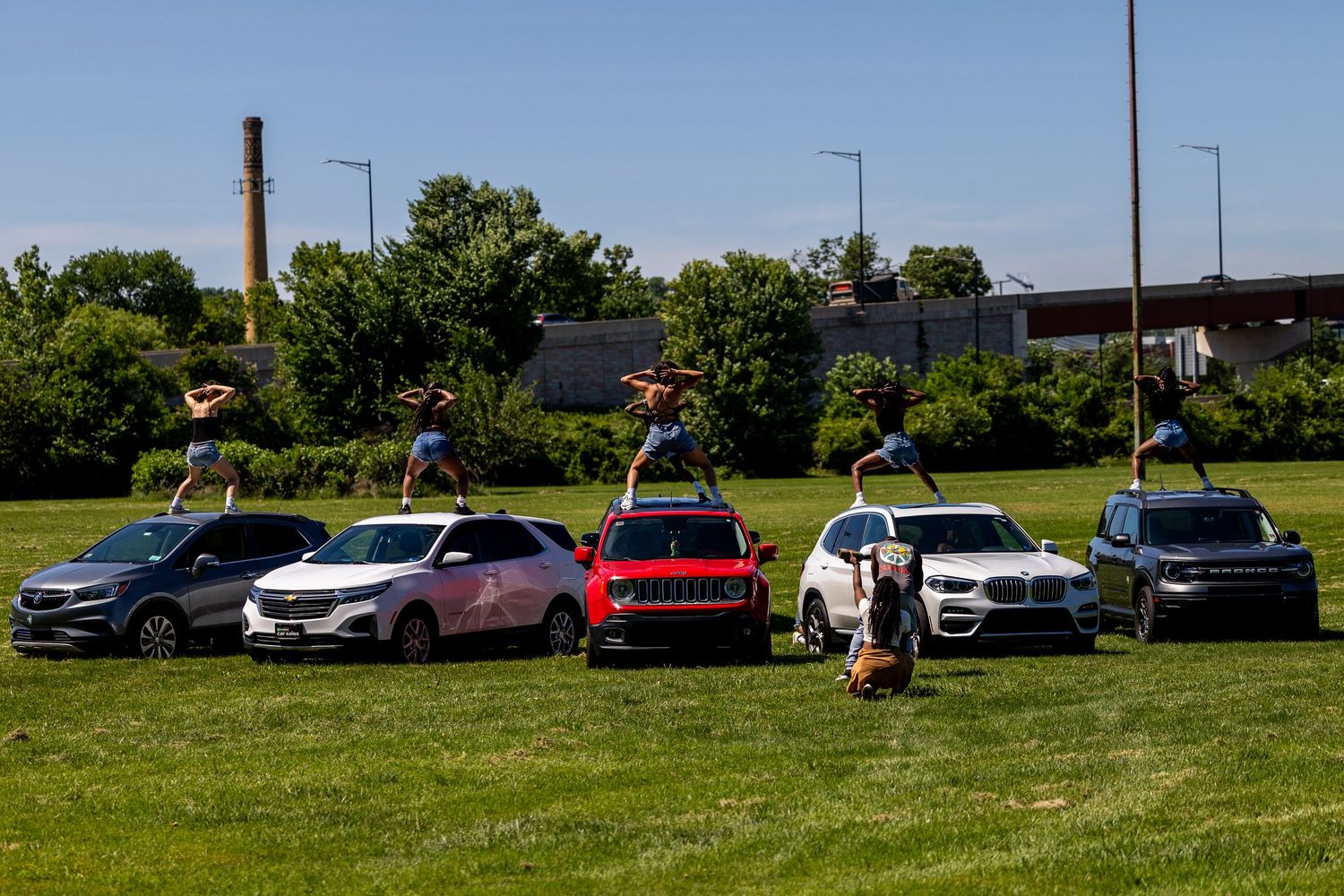 Acrobats perform balancing acts on top of colorful cars parked on a green lawn with trees in the background.