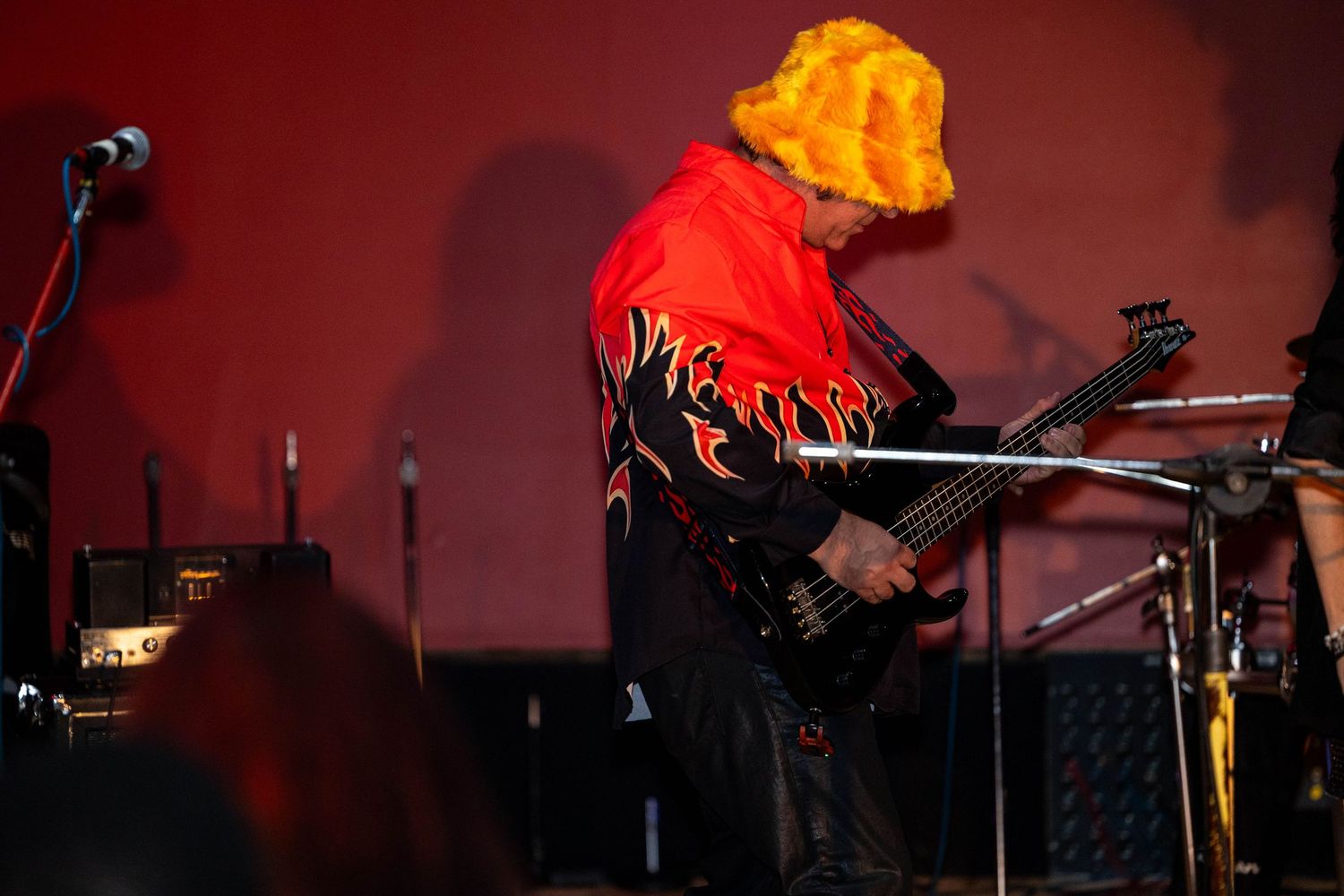 Guitarist in orange afro wig and flame-patterned jacket performs on stage under red lighting.
