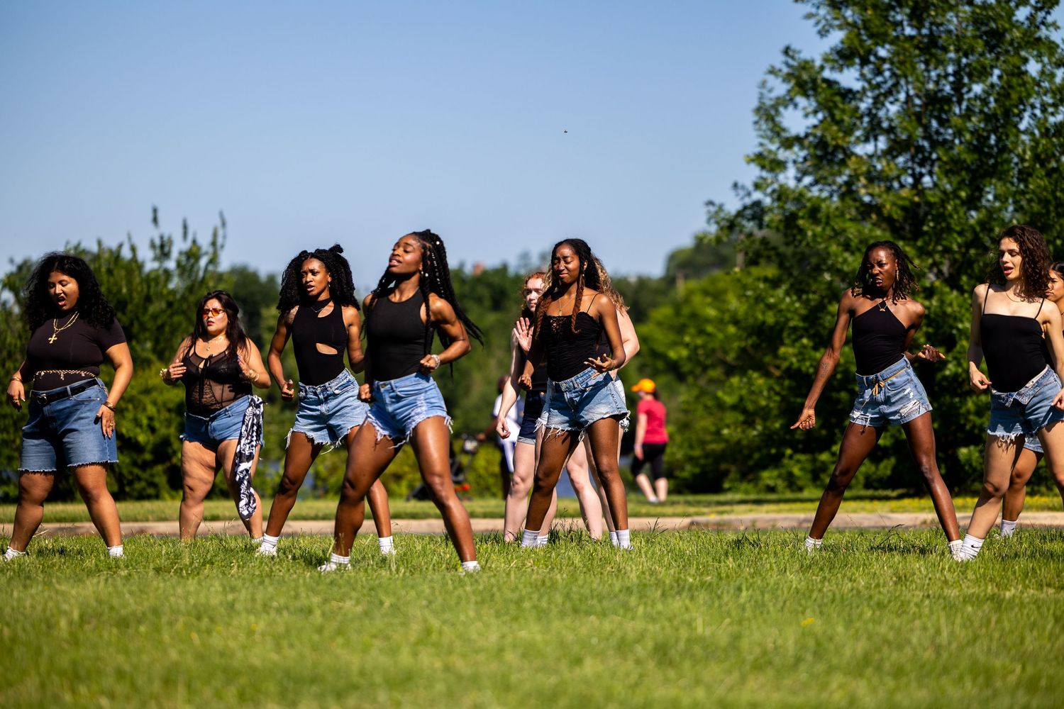 Group of young women dancing outdoors on green grass in summer, wearing denim shorts and black tops.
