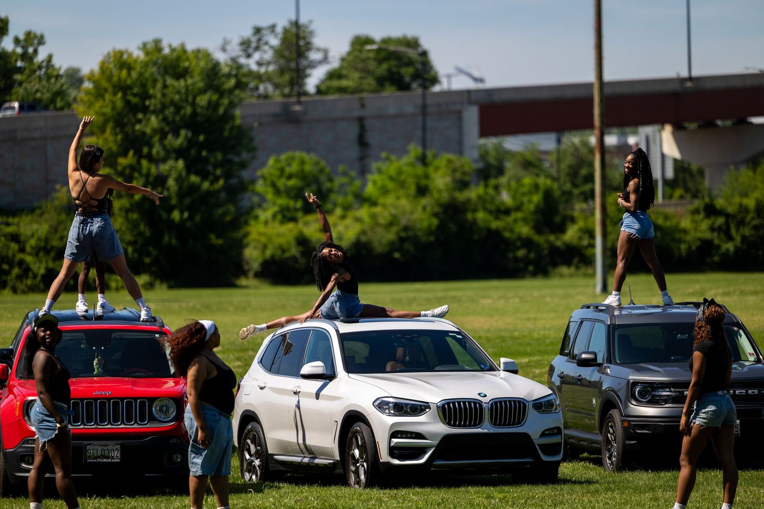 People standing on top of cars including a white BMW and red Jeep on a sunny day in a grassy field.