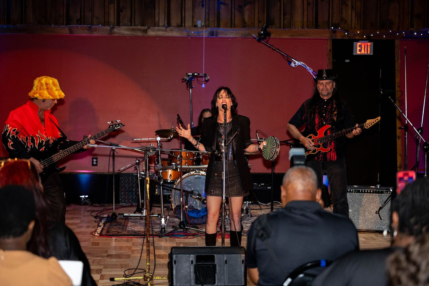 Live band performing on stage with female vocalist, guitarists, and drummer in a rustic indoor venue.