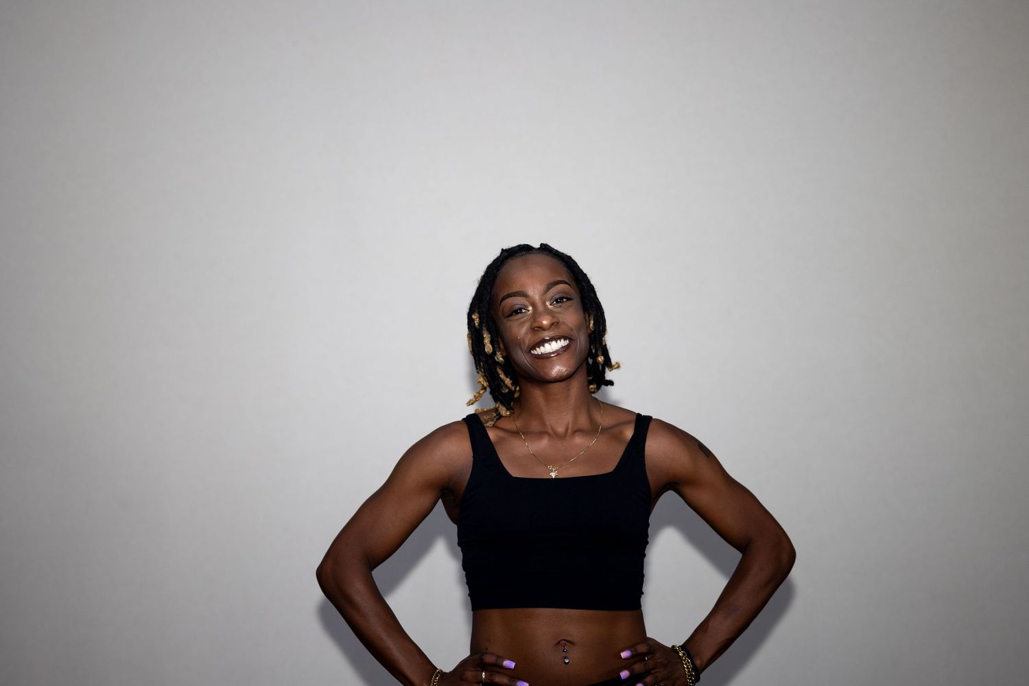 A fit Black woman in a black crop top laughs joyfully with hands on hips against white backdrop.