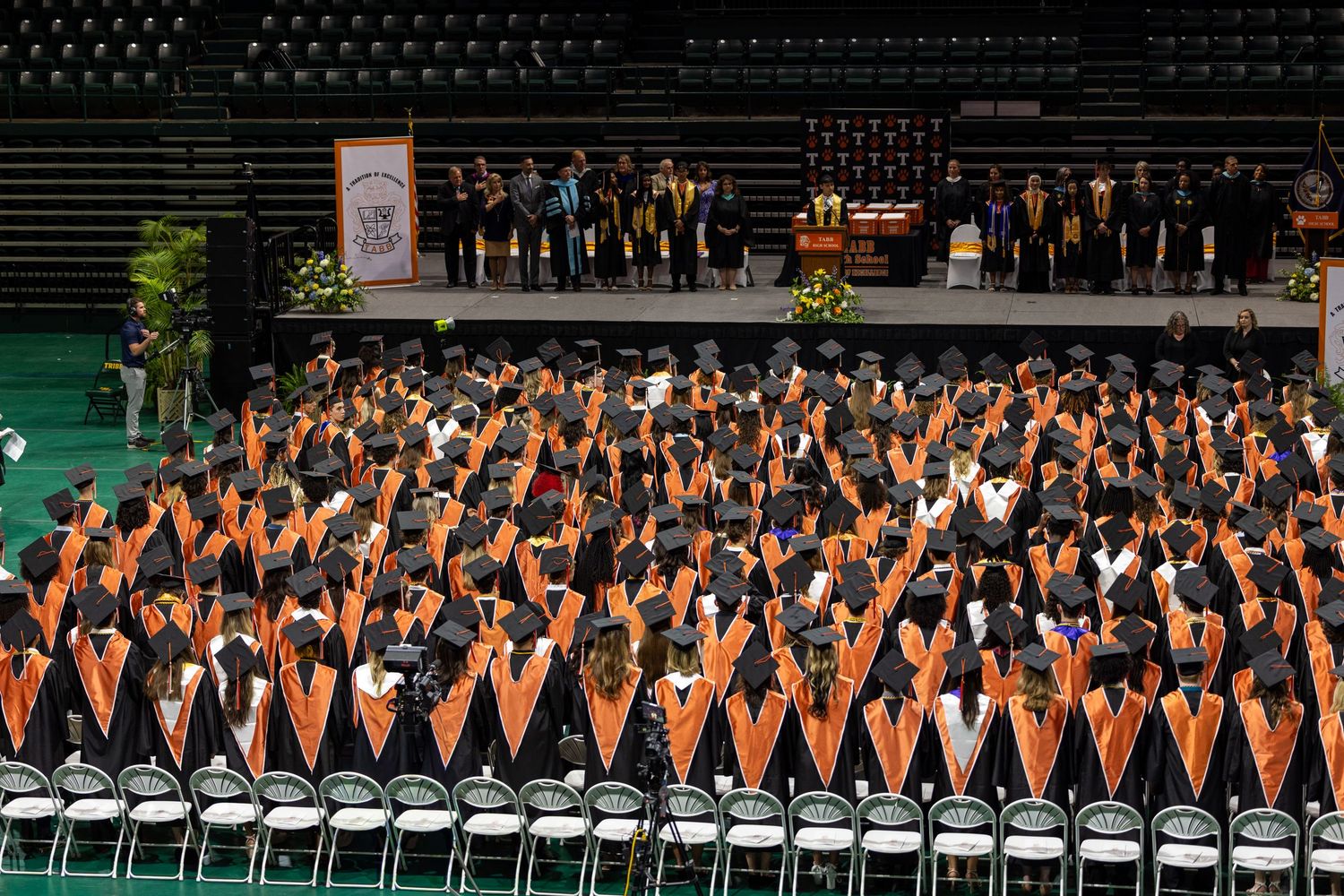 Large graduation ceremony with students in black gowns and orange stoles seated in rows before a stage.