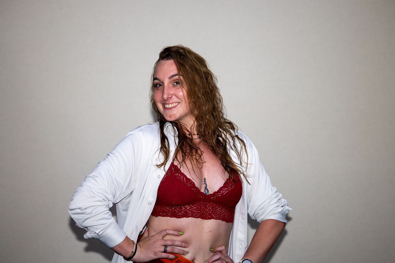 Smiling woman with curly hair wearing a red lace bralette and open white shirt, posing confidently indoors.
