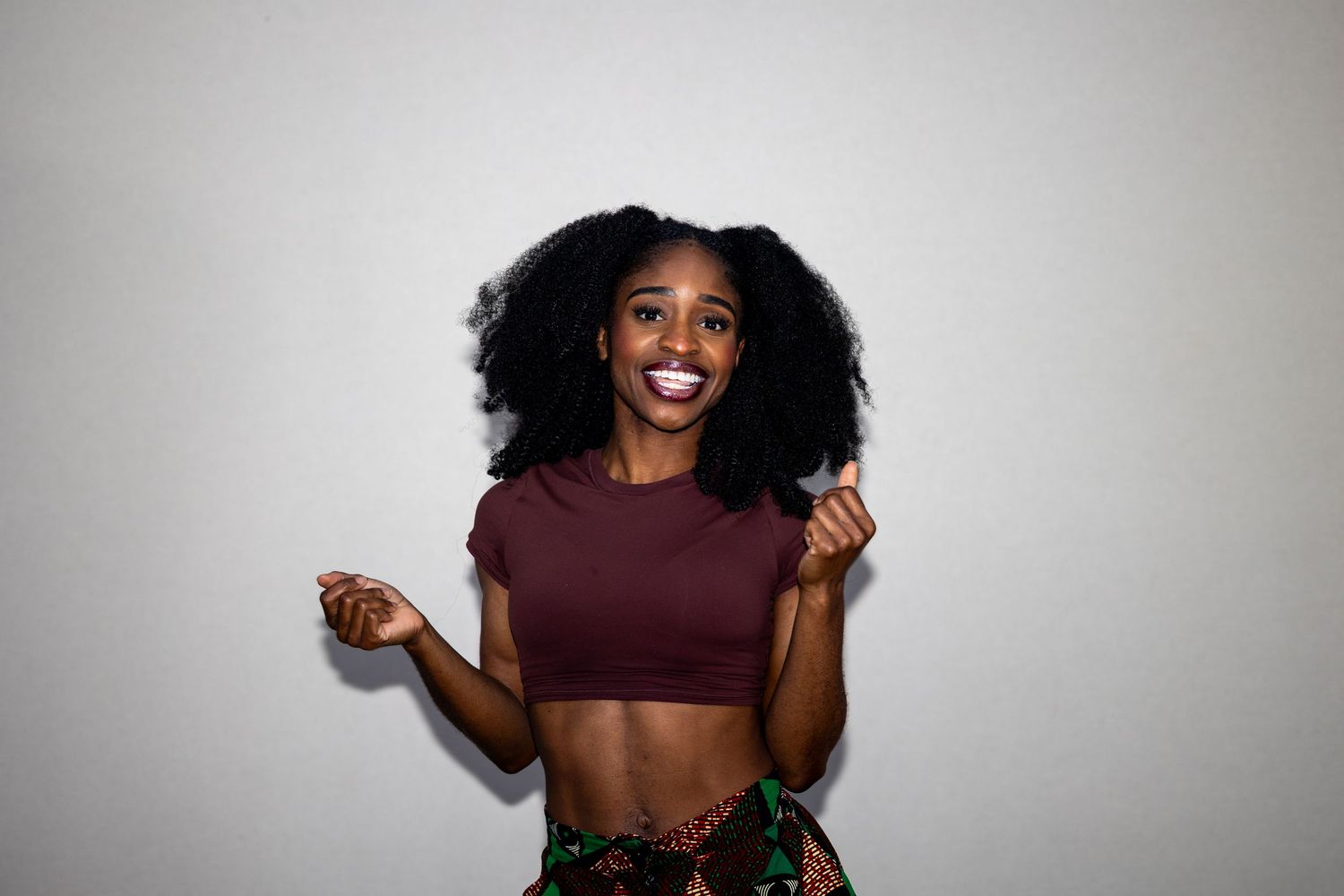 Joyful woman in maroon crop top dancing with arms outstretched against white background.