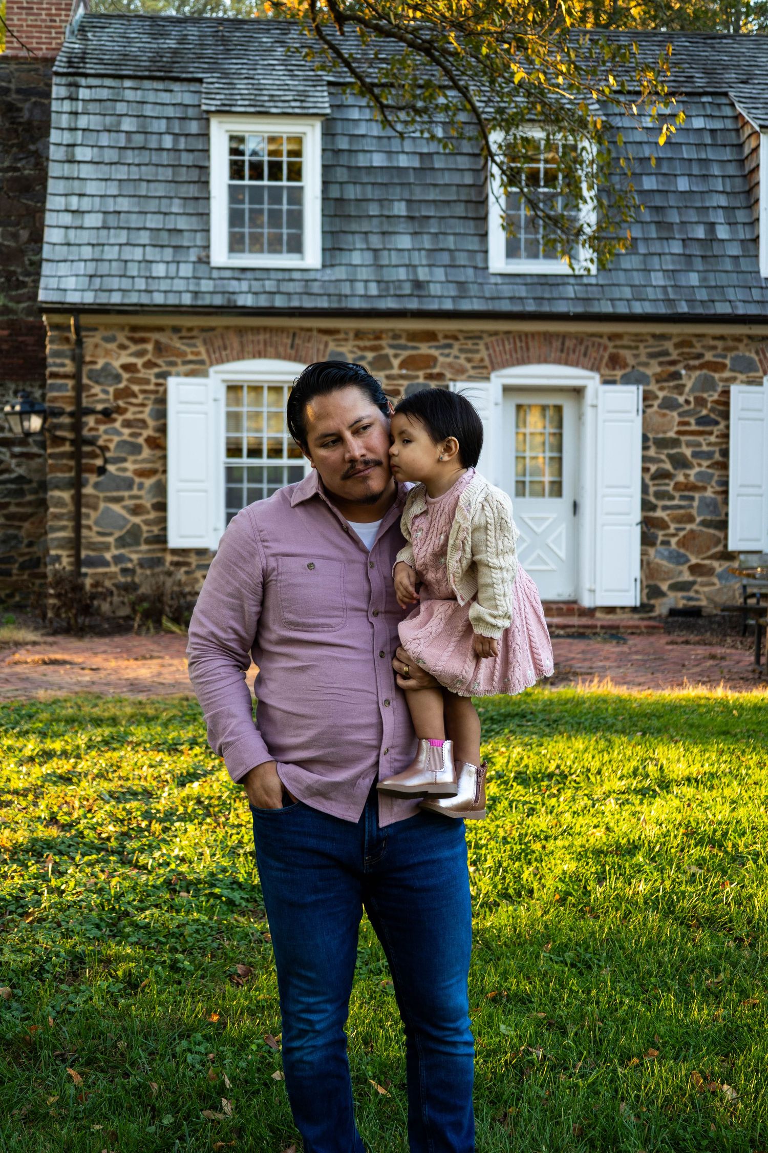 A man holds a young girl in a pink dress outside a stone house with white shutters on a sunny day.