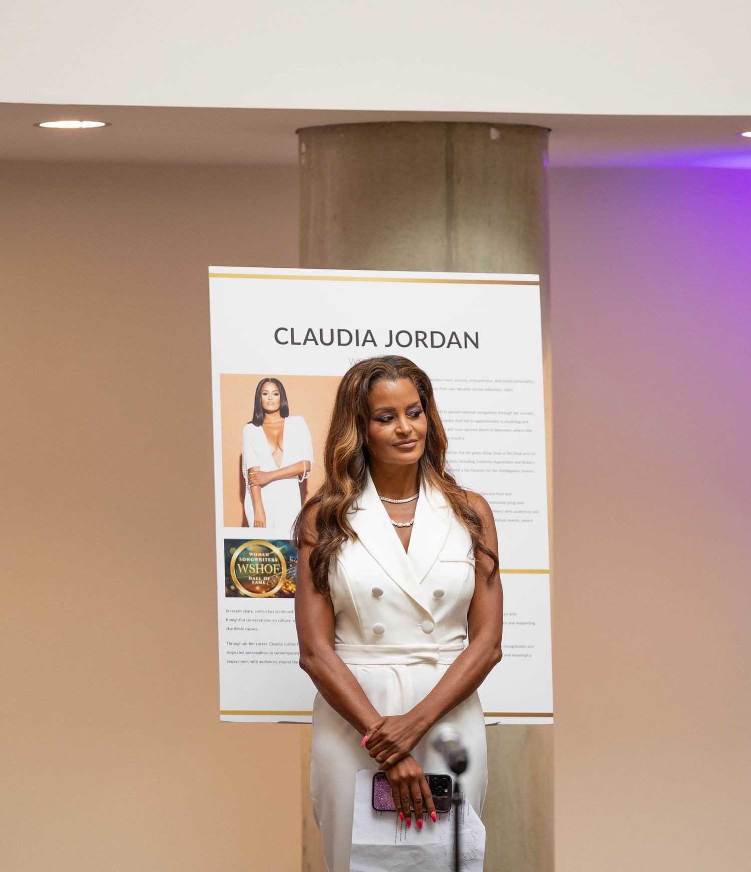 Woman in white blazer dress stands before Claudia Jordan biography display at formal recognition event.