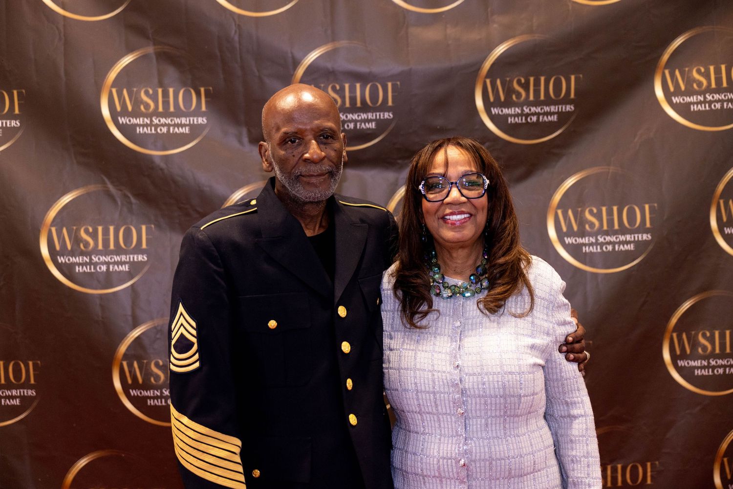 Man in military uniform and woman pose together at WSHOI Women Songwriters Hall of Fame event backdrop.