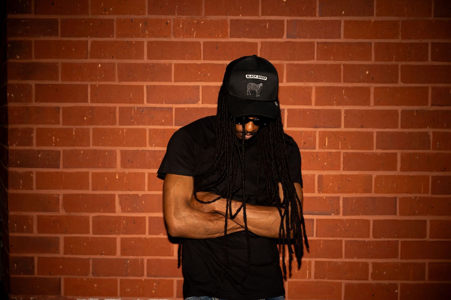 A muscular man with dreadlocks wearing a black cap and t-shirt poses against a red brick wall.