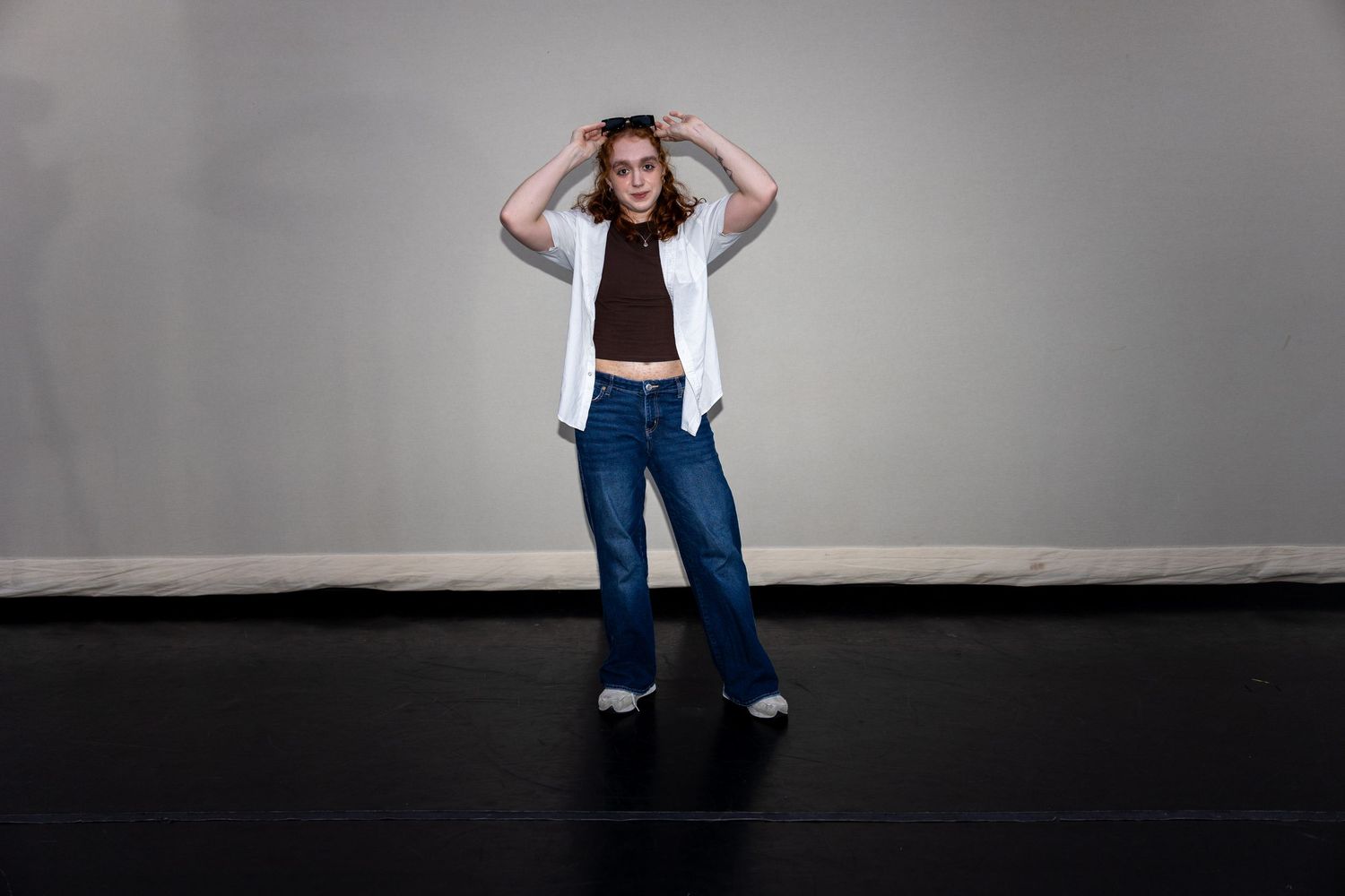A young woman in a white jacket, brown crop top, and wide-leg jeans stands with hands on head on a dark stage.