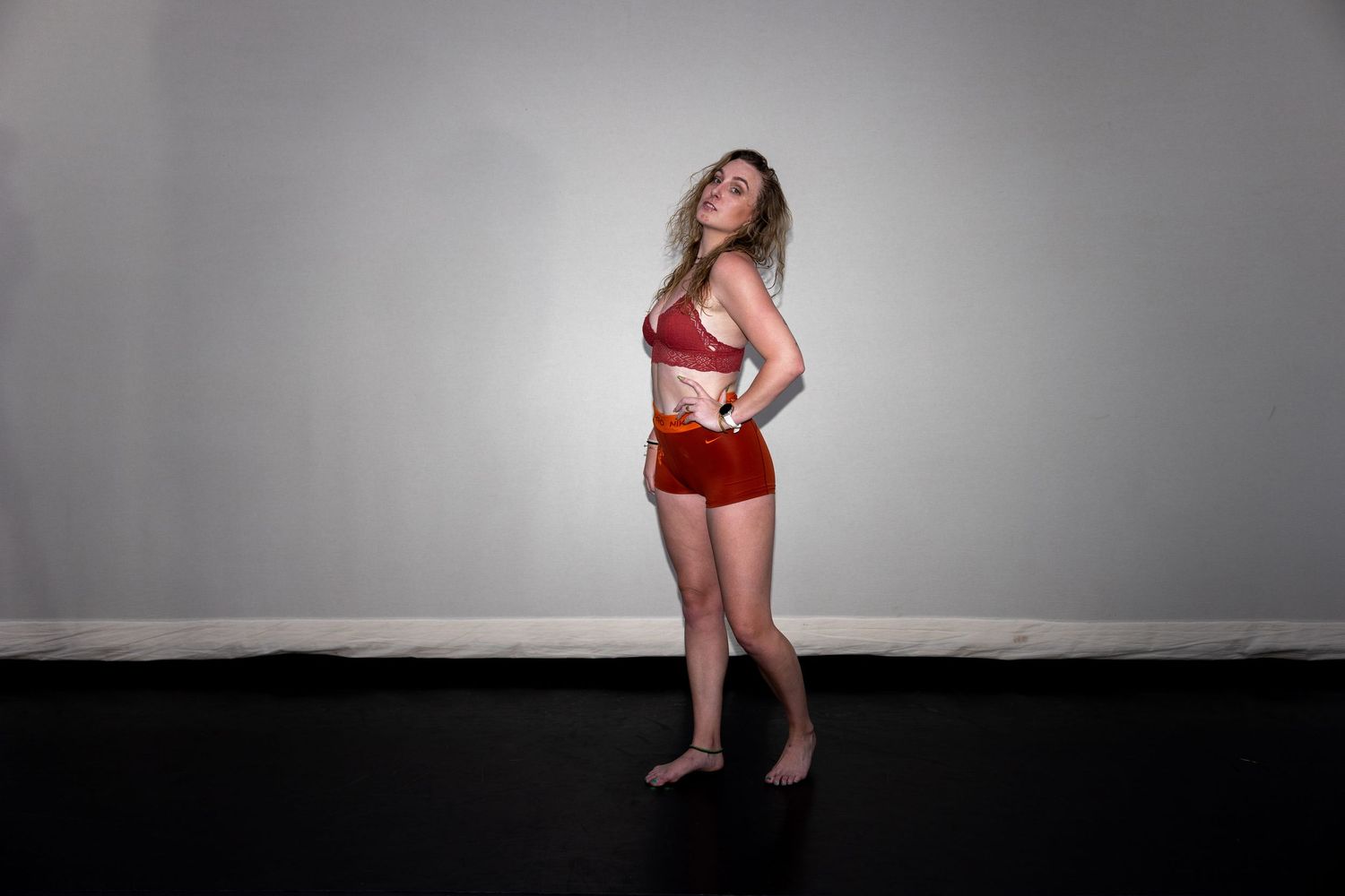 A woman in red sports bra and shorts poses sideways against a neutral gray studio background.