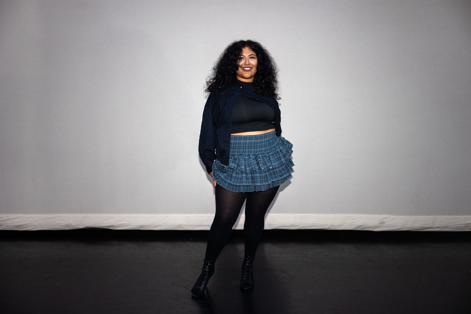 A woman in black crop top and blue plaid ruffled mini skirt stands confidently in a studio.